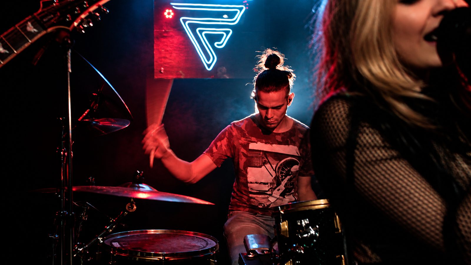 Baterista sob a placa neon com a vocalista em primeiro plano