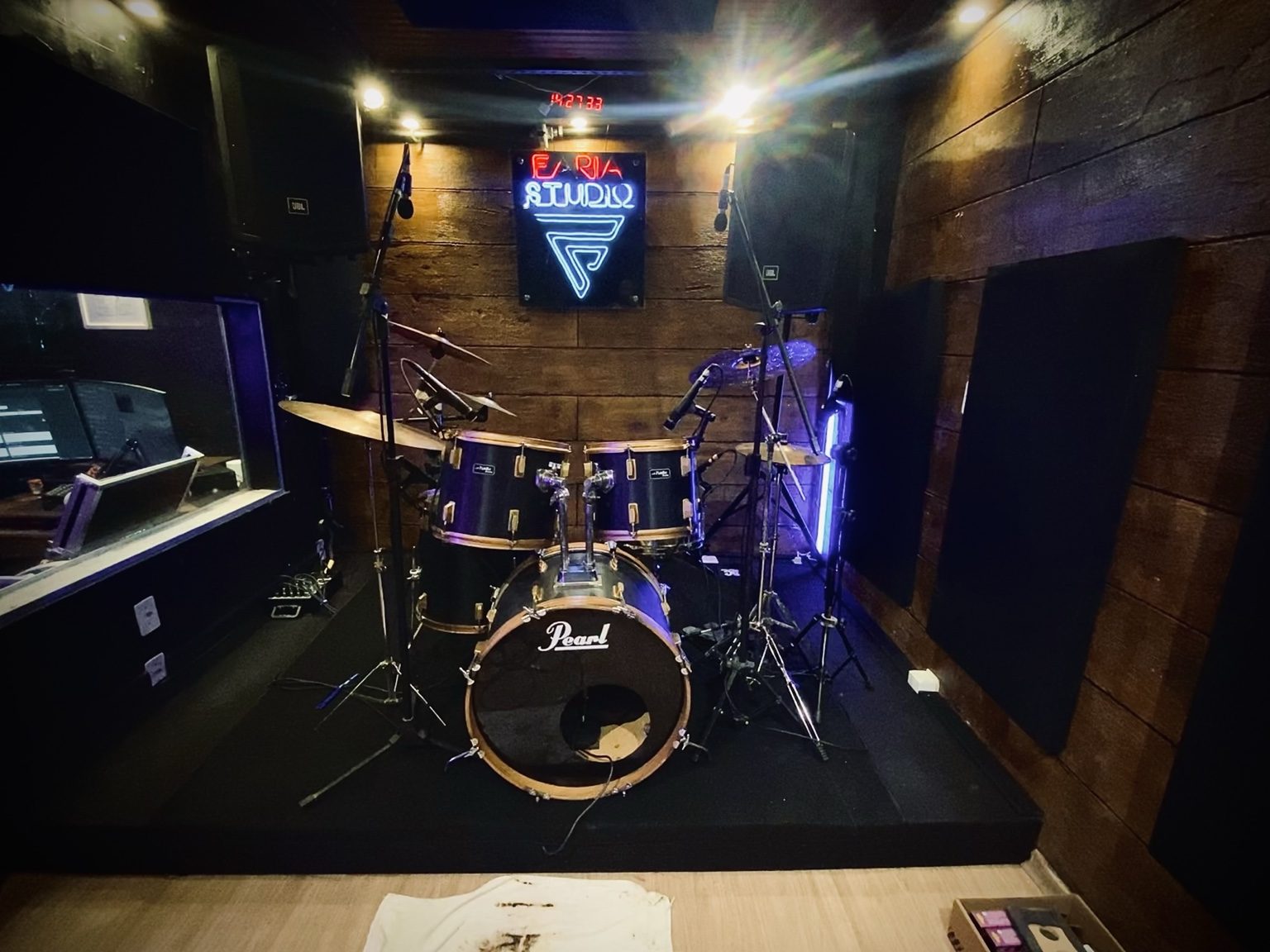 Bateria Pearl montada no palco da sala de ensaio, com a placa neon ao fundo